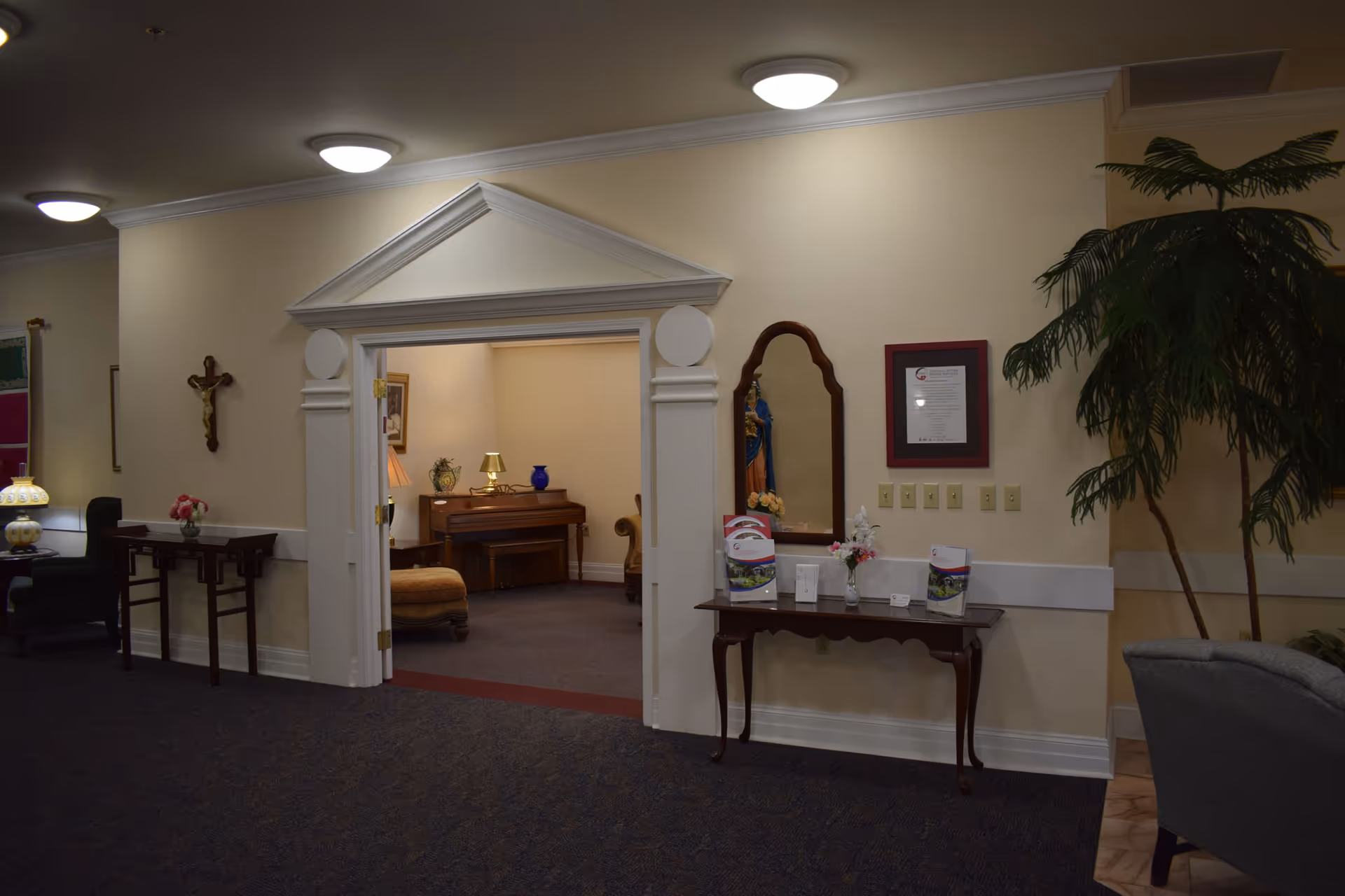 Interior view of a senior living facility showing a hallway with beige walls and dark carpet. There is a decorative doorway leading to a room with a piano, lamp, and cushioned chair. On the left wall, there is a crucifix and a small table with a flower vase. On the right wall, there is a mirror above a wooden table holding brochures and a small flower arrangement. A large potted plant is visible on the right side near a gray armchair.