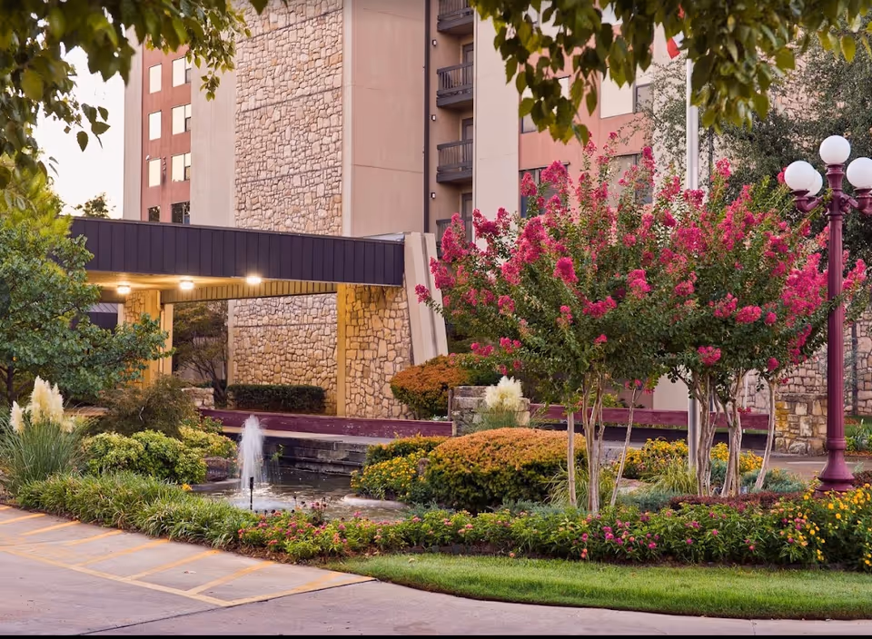 Exterior view of Senior Star at Burgundy Place showing a landscaped garden with flowering bushes, a small water fountain, and a multi-story building with stone and stucco facade in the background. There is a covered entrance with lights and a lamppost with globe lights on the right side.