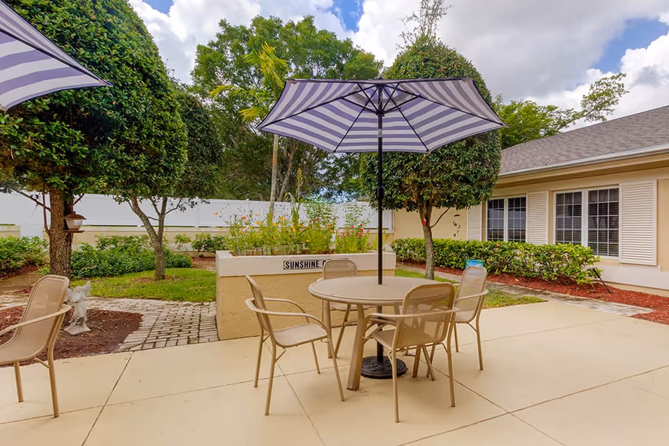 Outdoor patio area with a round table and four chairs under a purple and white striped umbrella. The patio is surrounded by neatly trimmed bushes and trees, with a raised garden bed labeled 'Sunshine Garden'. The building with windows and beige walls is visible in the background under a partly cloudy sky.