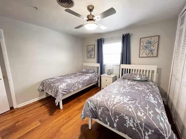 A bedroom with two single beds featuring white frames and matching gray floral bedspreads. Between the beds is a small white nightstand with a lamp. The room has wooden flooring, a ceiling fan with light, a window with dark curtains, and two framed floral artworks on the walls.