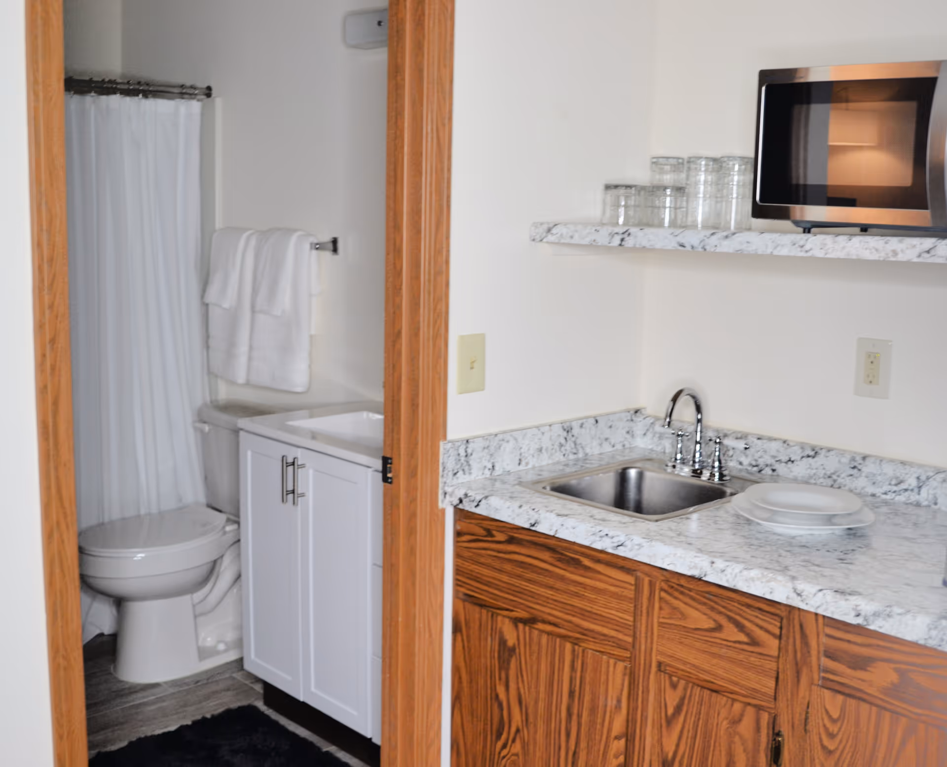 Small kitchenette with a sink, microwave, and marble-look countertop beside a doorway revealing a bathroom with a toilet and shower curtain.
