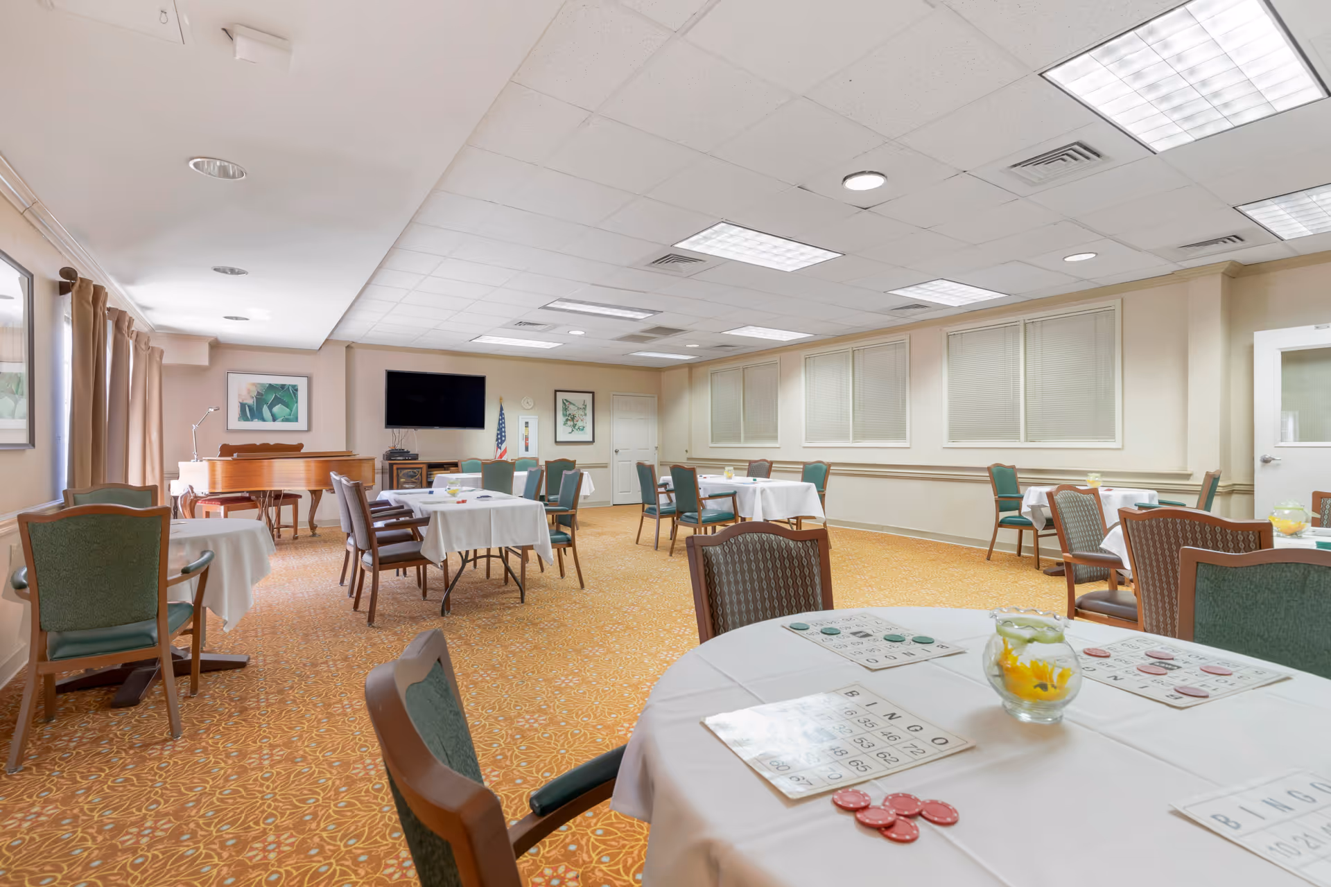 A senior living facility common room with multiple round tables covered with white tablecloths and green cushioned chairs. Bingo cards and chips are placed on the tables. The room has beige walls, a patterned carpet, a piano, a flat-screen TV mounted on the wall, and framed artwork. Windows with closed blinds line one wall.