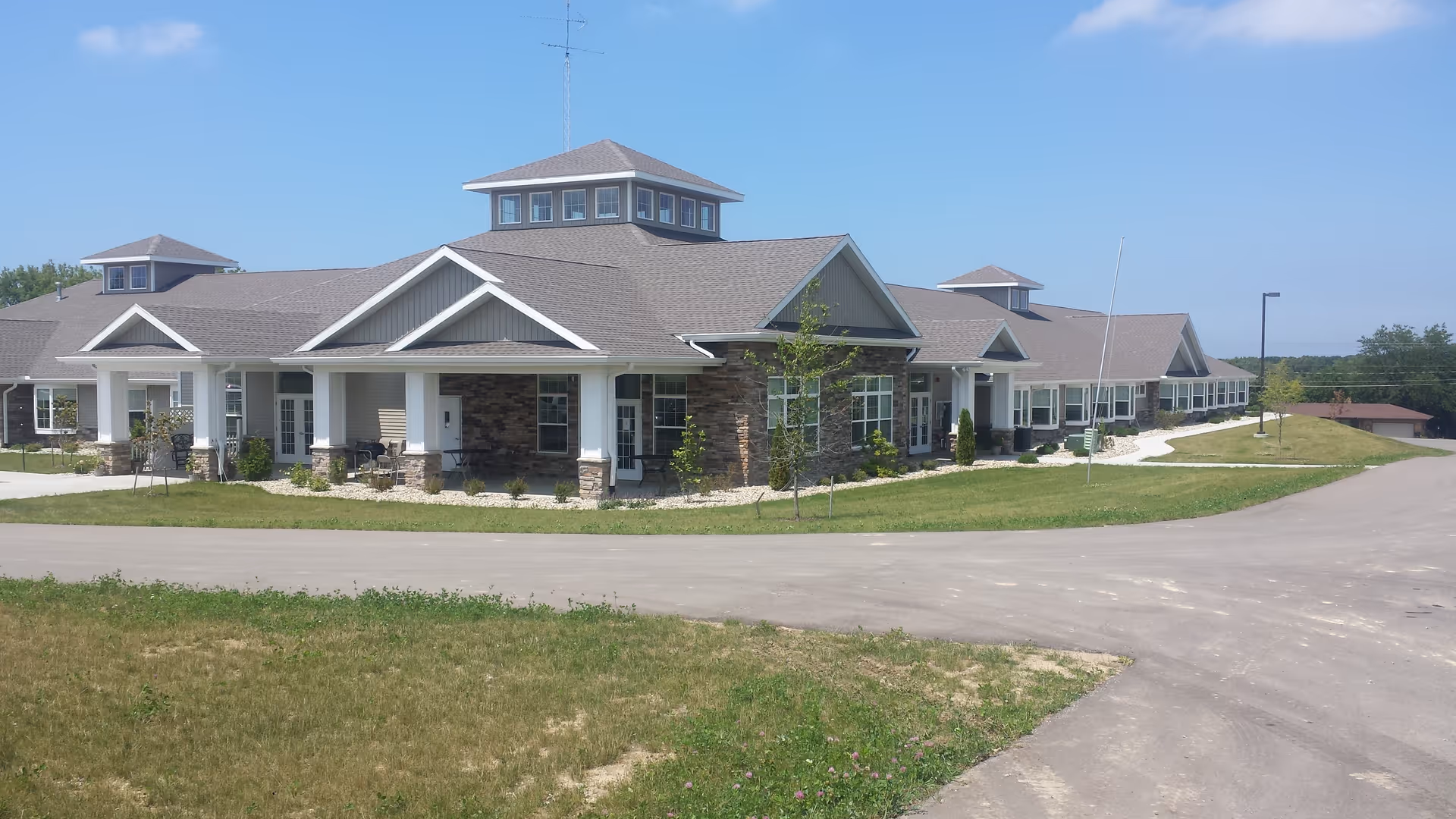 Exterior view of a single-story senior living facility building with a stone and siding facade, multiple windows, and a covered porch area. The building is surrounded by a paved driveway and grassy areas under a clear blue sky.