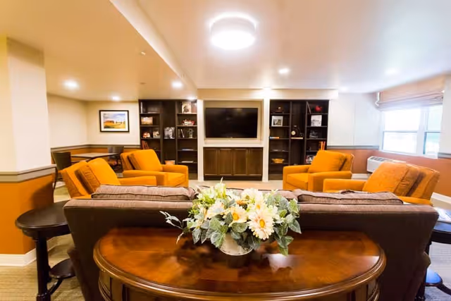 A cozy living room area with four orange armchairs arranged around a central coffee table, facing a large flat-screen TV mounted on a wall with built-in dark wood shelving units. A brown sofa with a floral arrangement on a wooden console table behind it is in the foreground. The room has warm lighting, beige walls with an orange accent, and a window with blinds on the right side.