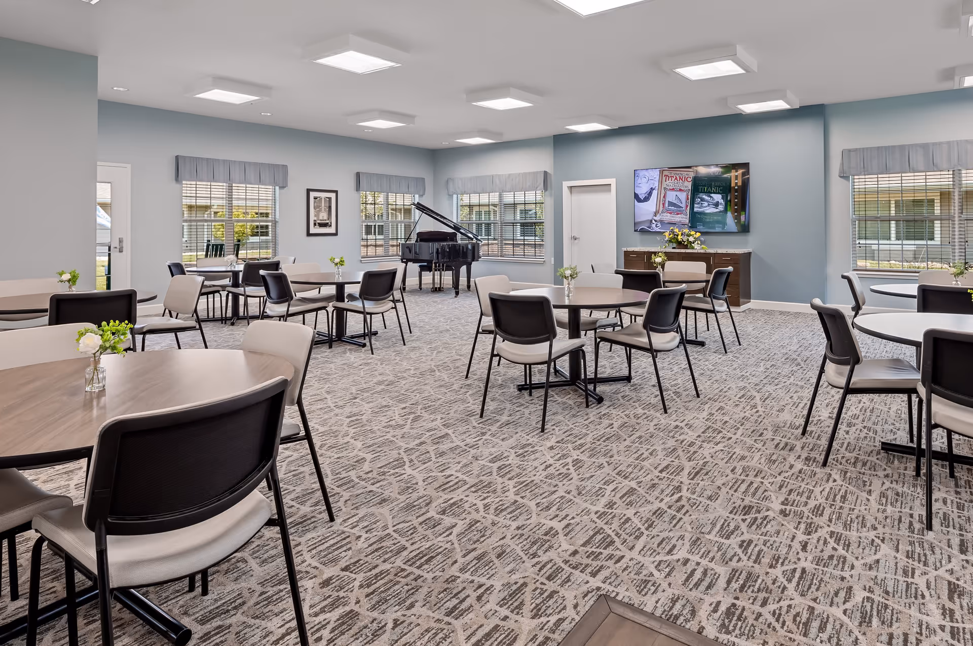 A spacious, well-lit common area with multiple round tables and chairs arranged throughout the room. The room features large windows with blinds and valances, a patterned carpet, and a grand piano in the corner. A flat-screen TV is mounted on the wall above a cabinet with flowers on top.
