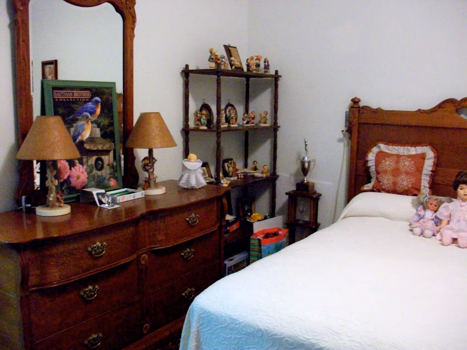 Cozy bedroom with a wooden dresser and mirror, shelving of knickknacks, and a bed with dolls.