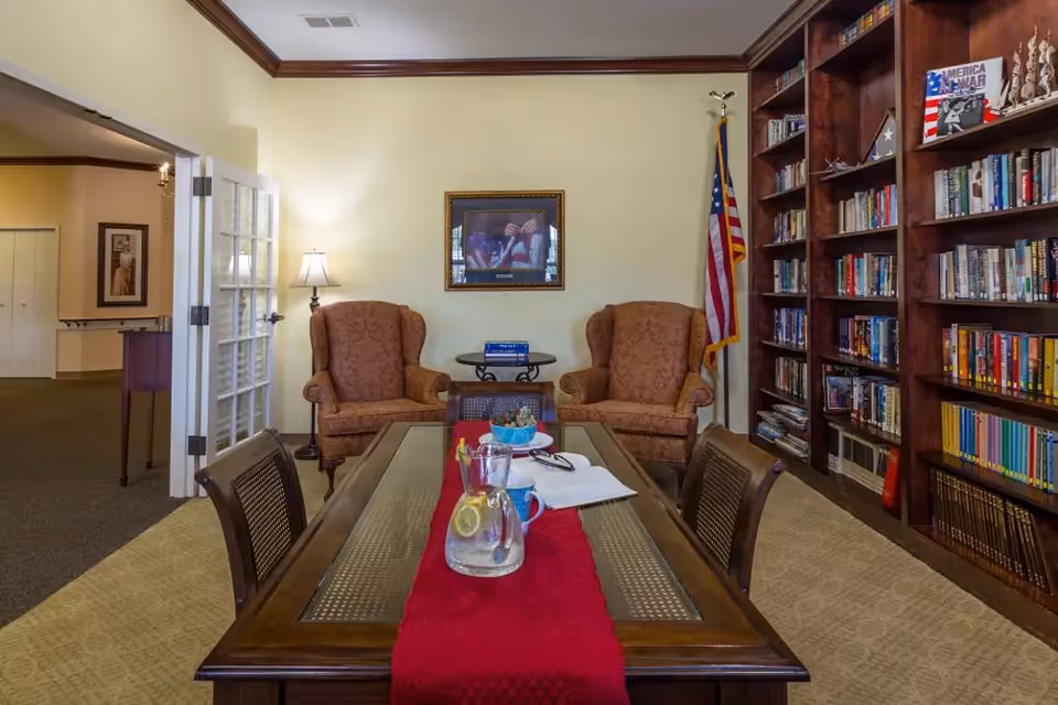 A cozy library-style sitting room with a long table topped by a red runner, two upholstered armchairs, and a wall of bookshelves.