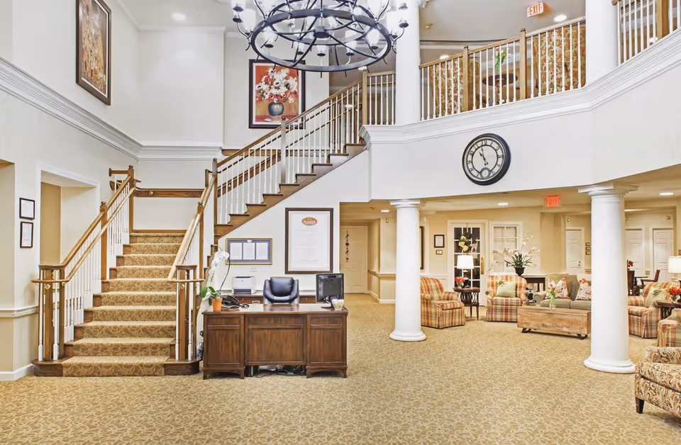 Interior view of a senior living facility lobby with a wooden reception desk, a staircase with carpeted steps and wooden railings, a large chandelier, a wall clock, and a seating area with patterned armchairs and sofas. The space is decorated with framed artwork and plants, and features beige carpeting and white columns.