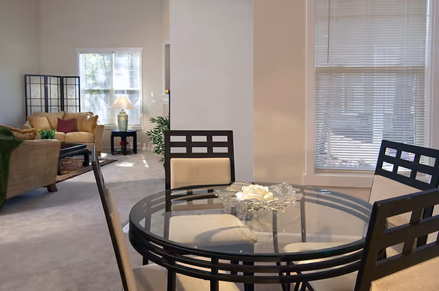Interior view of a senior living facility showing a glass-top dining table with four chairs in the foreground and a living room area with sofas, a side table with a lamp, and a window in the background.