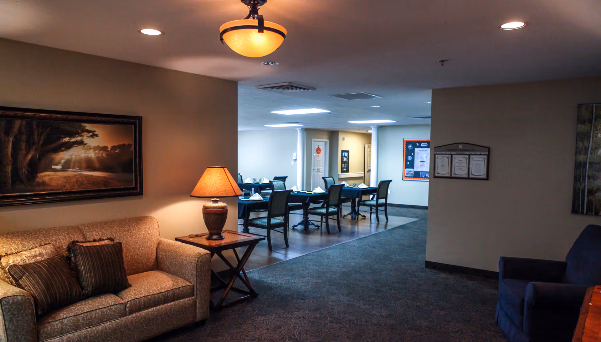 Interior view of a senior living facility showing a cozy sitting area with a beige couch, a side table with a lamp, and a framed picture on the wall. In the background, there is a dining area with tables and chairs set up for meals.