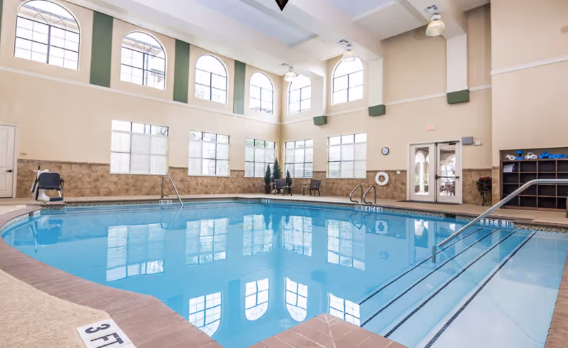 Indoor swimming pool area with clear blue water, surrounded by beige tiled walls and large windows with arched tops letting in natural light. There are pool stairs with handrails, a pool lift chair, chairs and tables along the far wall, and shelves with pool equipment. The ceiling is high with hanging lights.