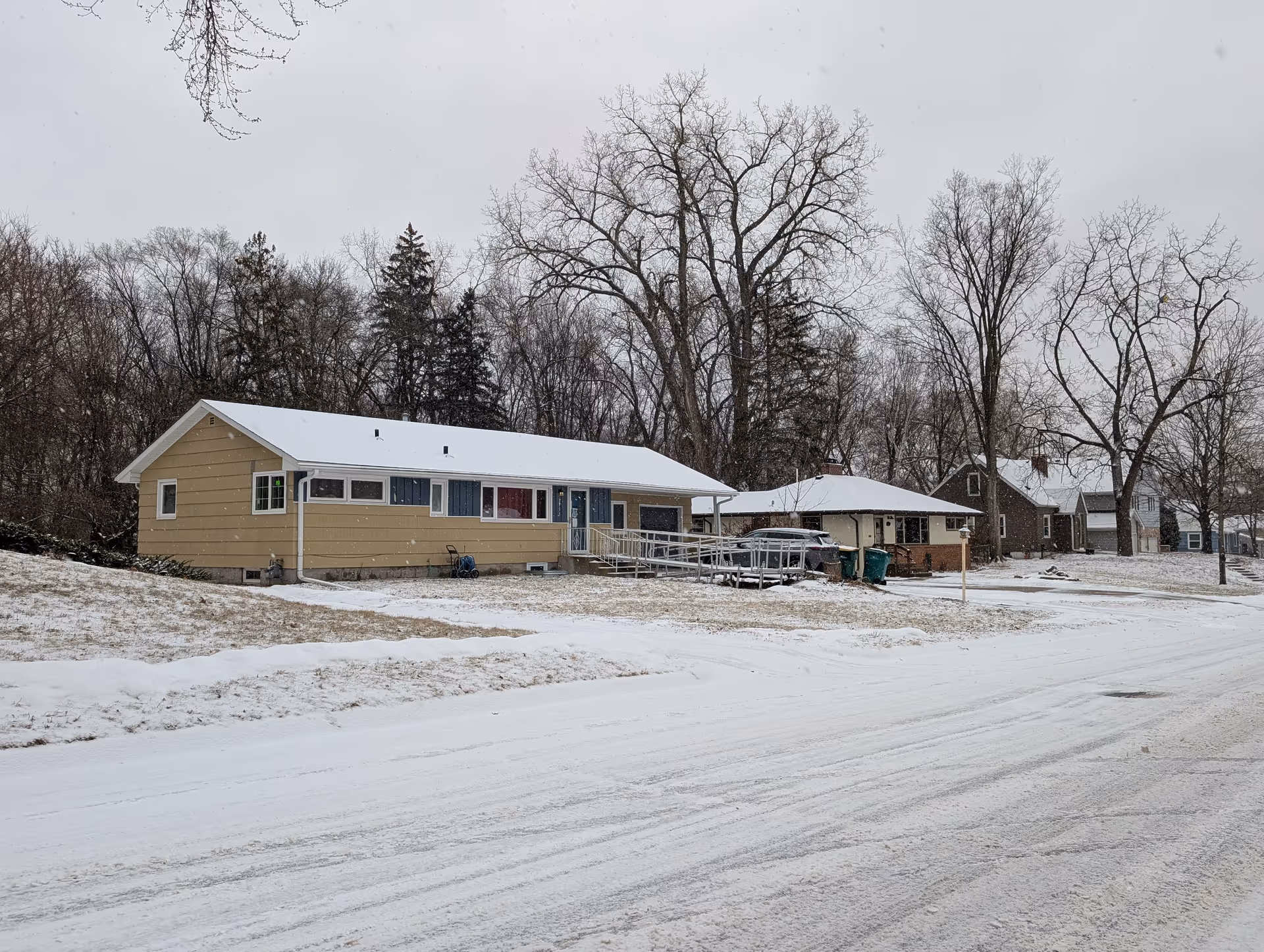 A snowy residential street with several single-story houses. The closest house is beige with a white roof covered in snow and has a wheelchair ramp leading to the front door. Leafless trees are visible in the background under a gray sky.