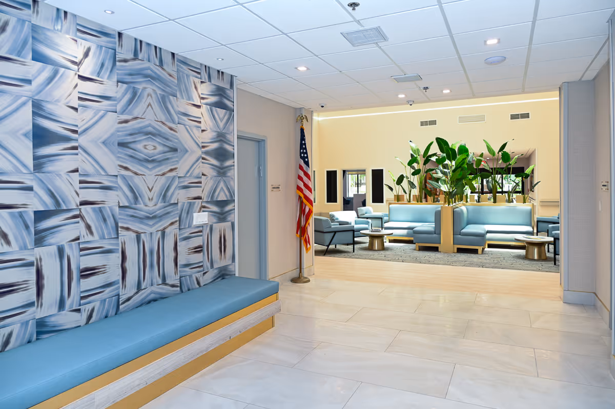 Interior view of a senior living facility lobby area featuring a blue cushioned bench along a wall with abstract blue and white patterned panels. An American flag stands near a doorway, and beyond the lobby is a seating area with light blue sofas, armchairs, small round tables, and several green plants. The space has a tiled floor and a drop ceiling with recessed lighting.