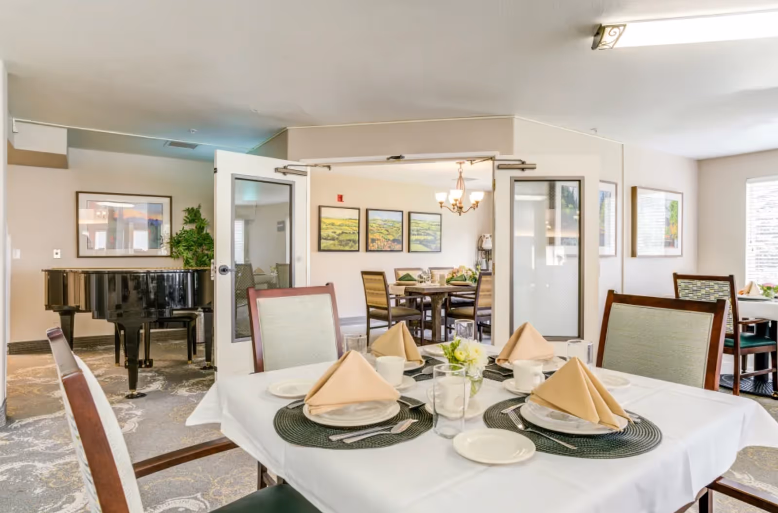 A dining area in a senior living facility with a table set for four, featuring beige napkins, plates, cups, and silverware on a white tablecloth. In the background, there is a black grand piano, framed artwork on the walls, and additional dining tables with chairs. The room is well-lit with natural light coming through windows.