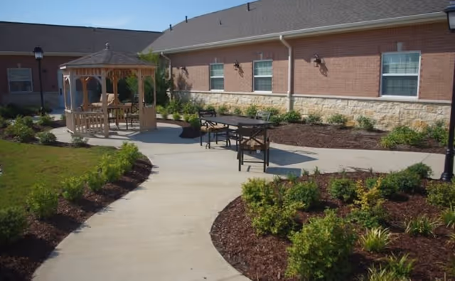 Outdoor courtyard area with a concrete pathway, landscaped garden beds with green shrubs, a wooden gazebo with seating, and a table with chairs. The building exterior is visible with brick and stone walls and several windows.