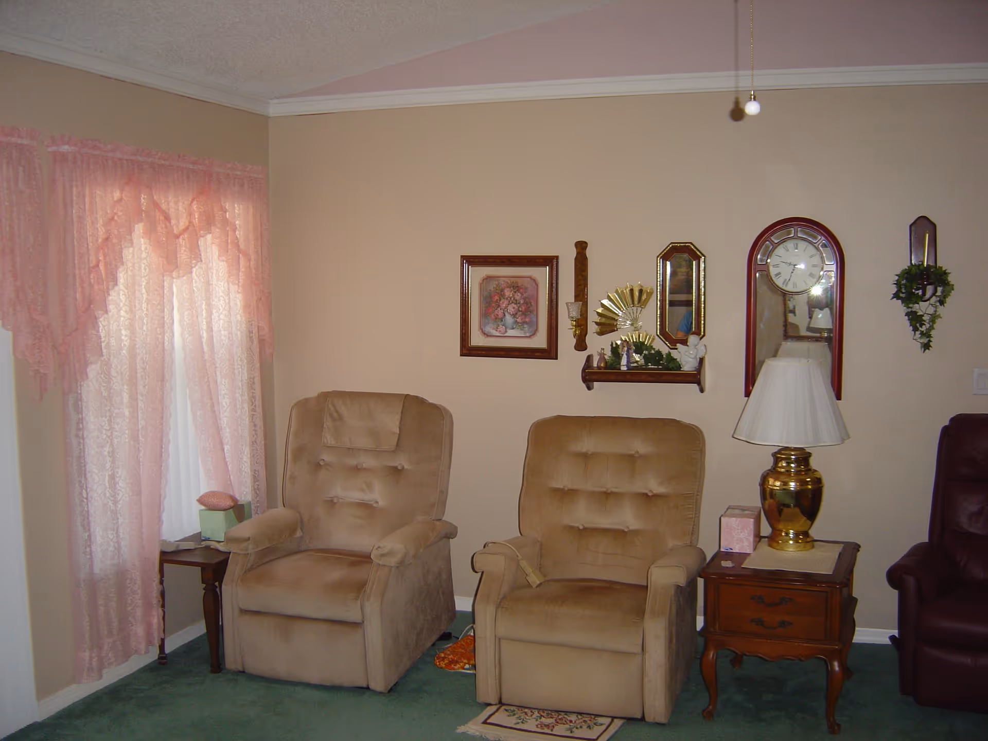 A cozy living room area with two beige recliner chairs placed side by side on a green carpet. To the left, there is a window with pink lace curtains and a small wooden side table with a tissue box. On the right side, there is a wooden side table with a brass lamp and a pink box. The wall behind the chairs is decorated with a framed floral painting, a small shelf holding decorative items, a wall clock with a pendulum, a small mirror, and a wall-mounted candle holder with greenery.