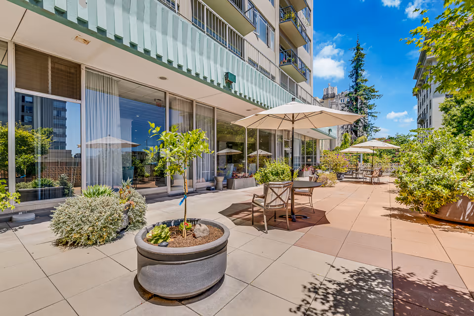 Sunlit outdoor patio at Lake Park Senior Living with tables and umbrellas, potted plants, and the building façade.