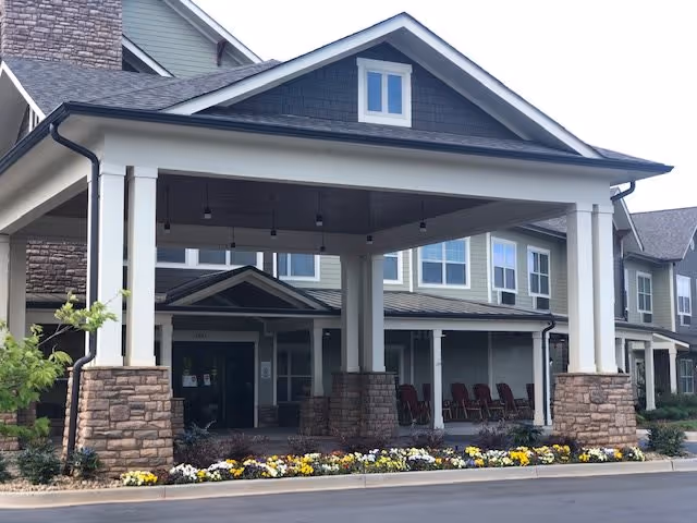 Front exterior view of a senior living facility with a covered entrance supported by white columns with stone bases. There are flower beds with colorful flowers along the driveway and a row of chairs on the porch area.
