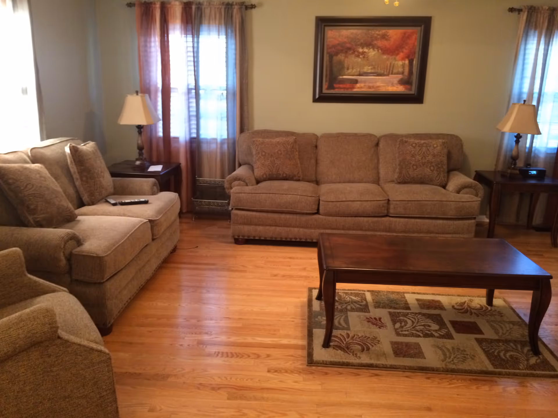A cozy living room with two beige sofas and a matching armchair arranged around a wooden coffee table on a patterned rug. There are two side tables with lamps on either side of the sofas, and a framed painting of a tree-lined path hangs on the wall. Light filters in through two windows with sheer curtains.