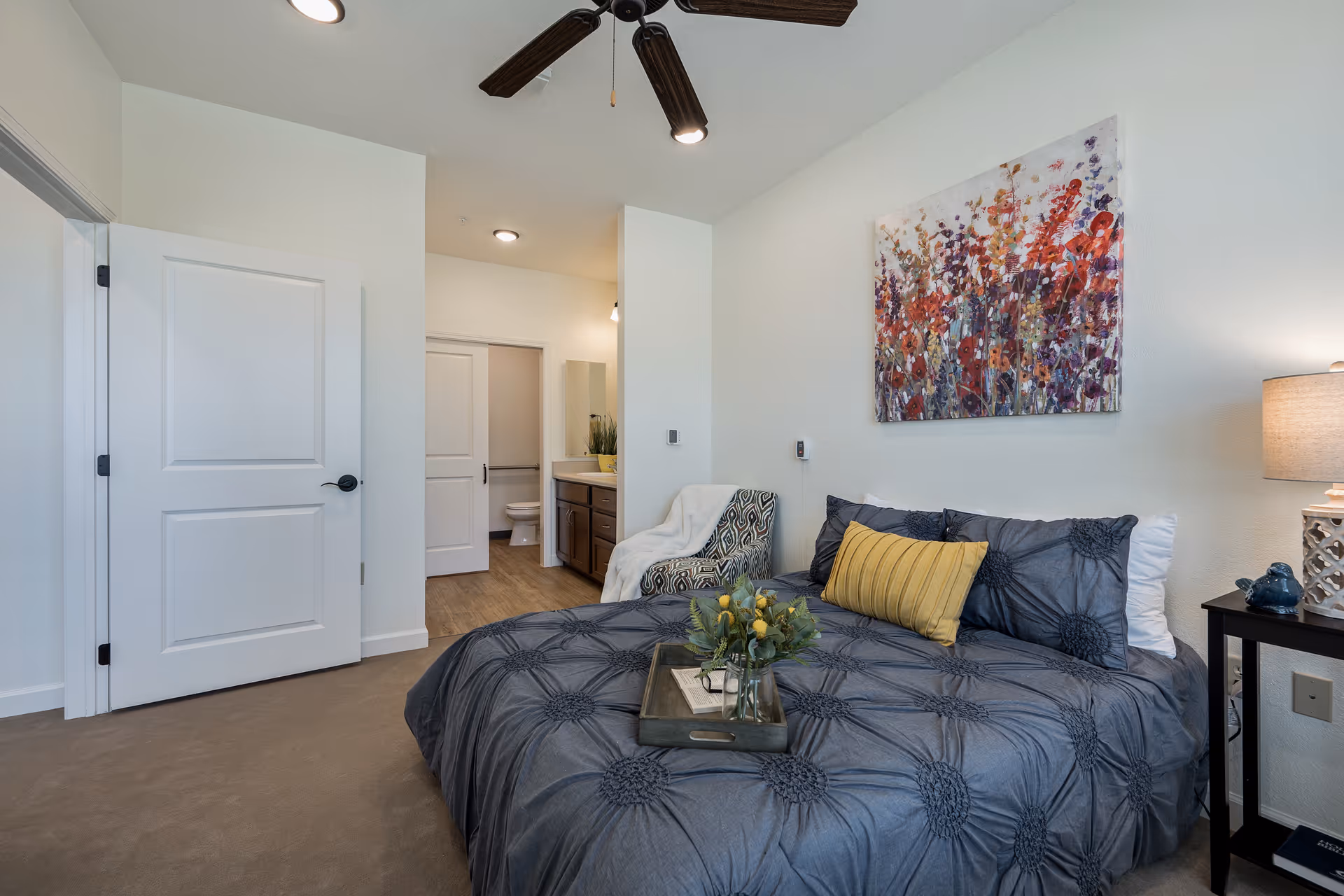Staged bedroom with a made bed, decorative pillows and artwork, a bedside table and lamp, and an open door leading to an en-suite bathroom.