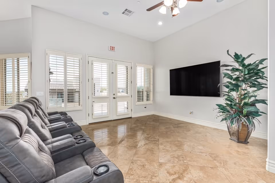 A bright and spacious living room with a row of black leather recliner chairs facing a large flat-screen TV mounted on a white wall. The room has beige tiled flooring, white walls, a ceiling fan with lights, and large windows with white shutters. A large potted plant is placed in the corner near the TV. Double glass doors with shutters lead outside.
