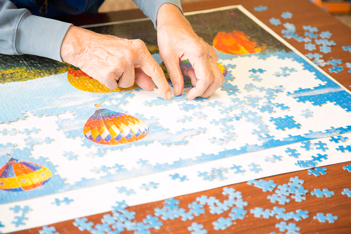 Close-up of elderly hands assembling a colorful hot air balloon jigsaw puzzle on a wooden table with scattered puzzle pieces.