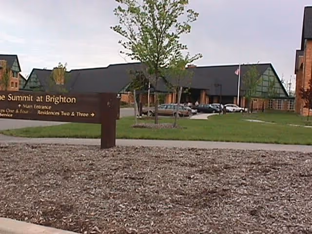 Exterior view of The Summit at Brighton Independent Living facility showing a large building with peaked roofs, several parked cars, a green lawn with young trees, and a brown sign in the foreground indicating directions to the main entrance and residences.