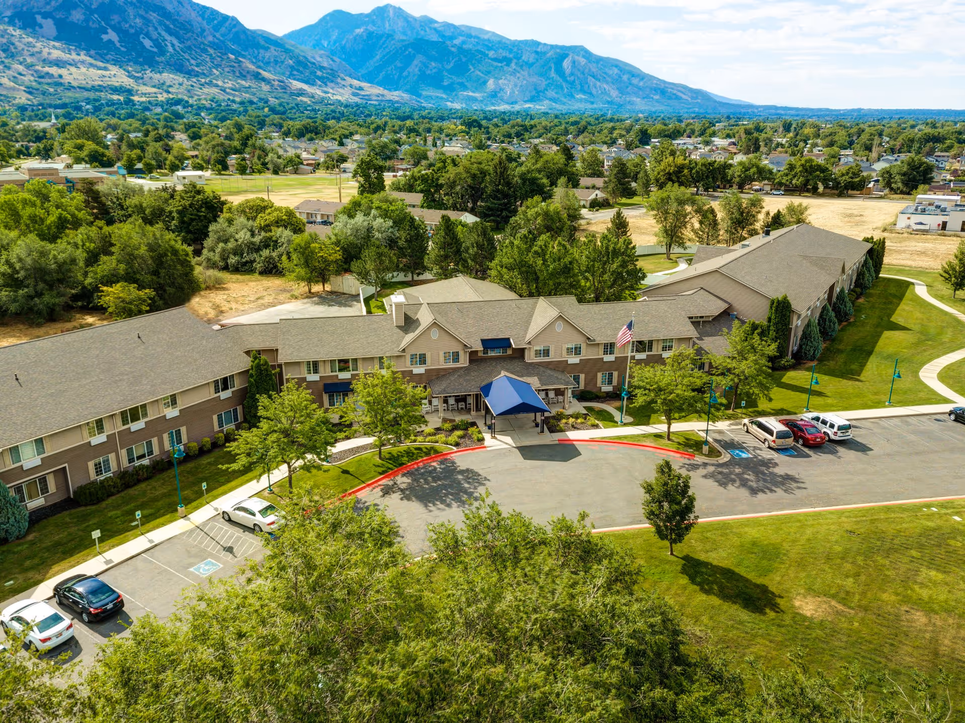 Aerial view of The Auberge at North Ogden senior living facility showing a large two-story building with a blue canopy entrance, surrounded by trees, parking spaces, and green lawns, with mountains visible in the background.