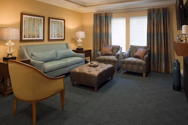 A cozy living room with a light blue sofa, two patterned armchairs with brown pillows, a beige armchair, and a tufted ottoman in the center. There are two framed pictures of trees on the wall above the sofa, two table lamps on dark wooden side tables, and large windows with striped curtains letting in natural light.