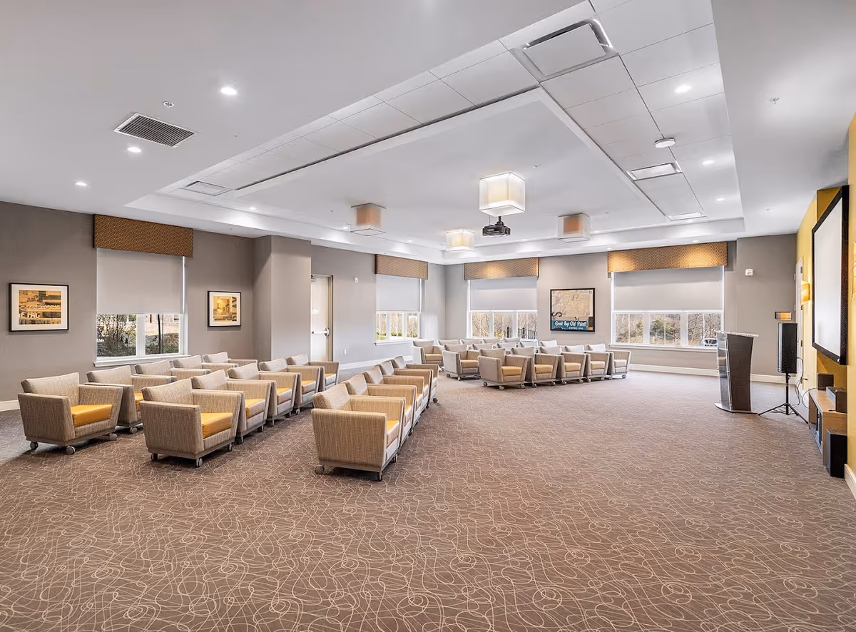 A spacious, well-lit meeting or presentation room with rows of beige armchairs arranged facing a large screen and podium. The room has large windows with blinds, framed artwork on the walls, and a patterned carpet.