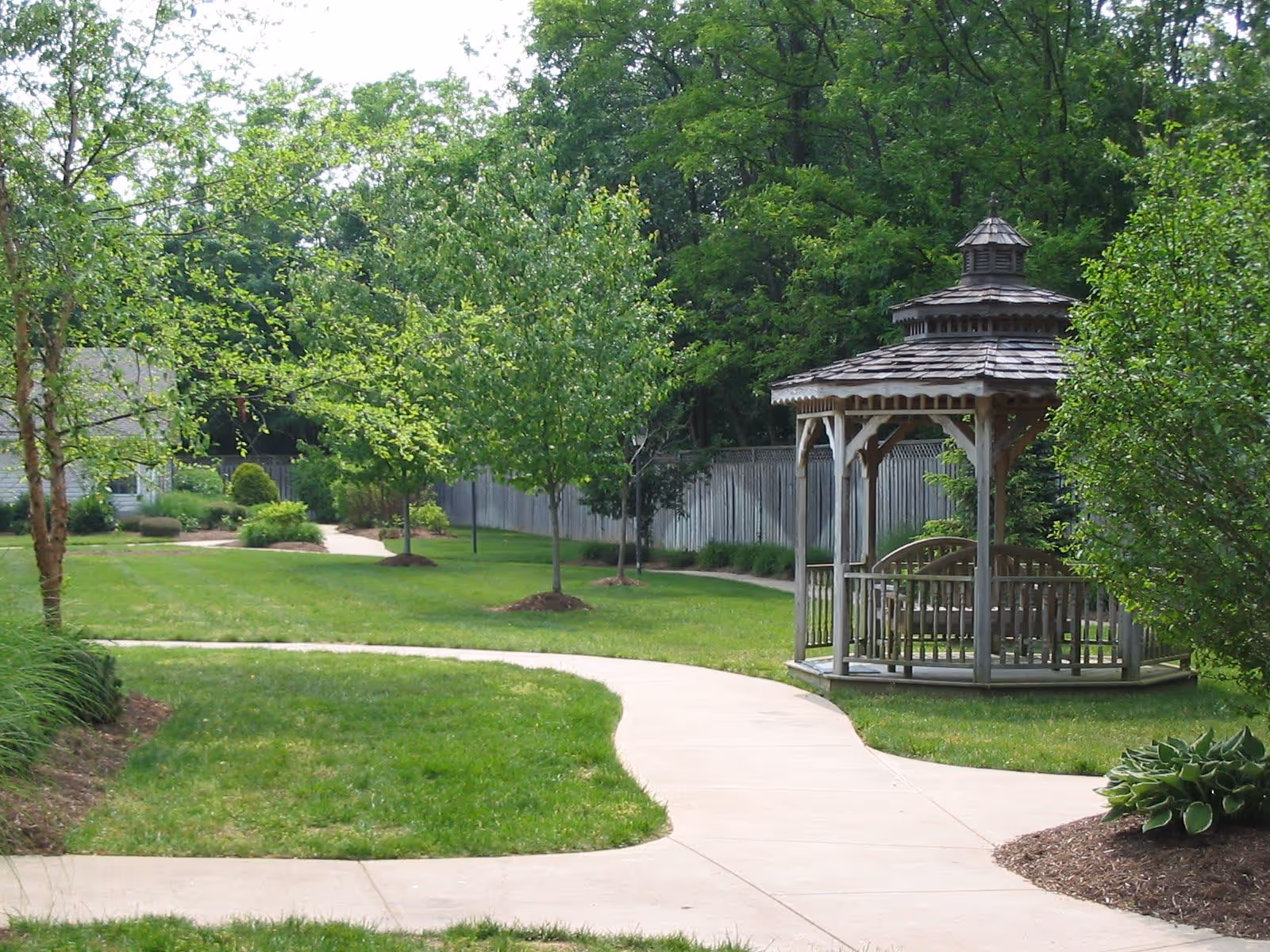 A winding concrete pathway through a well-maintained green lawn with small trees and shrubs. To the right of the path is a wooden gazebo with a shingled roof and a bench inside. In the background, there is a wooden fence and dense green trees.