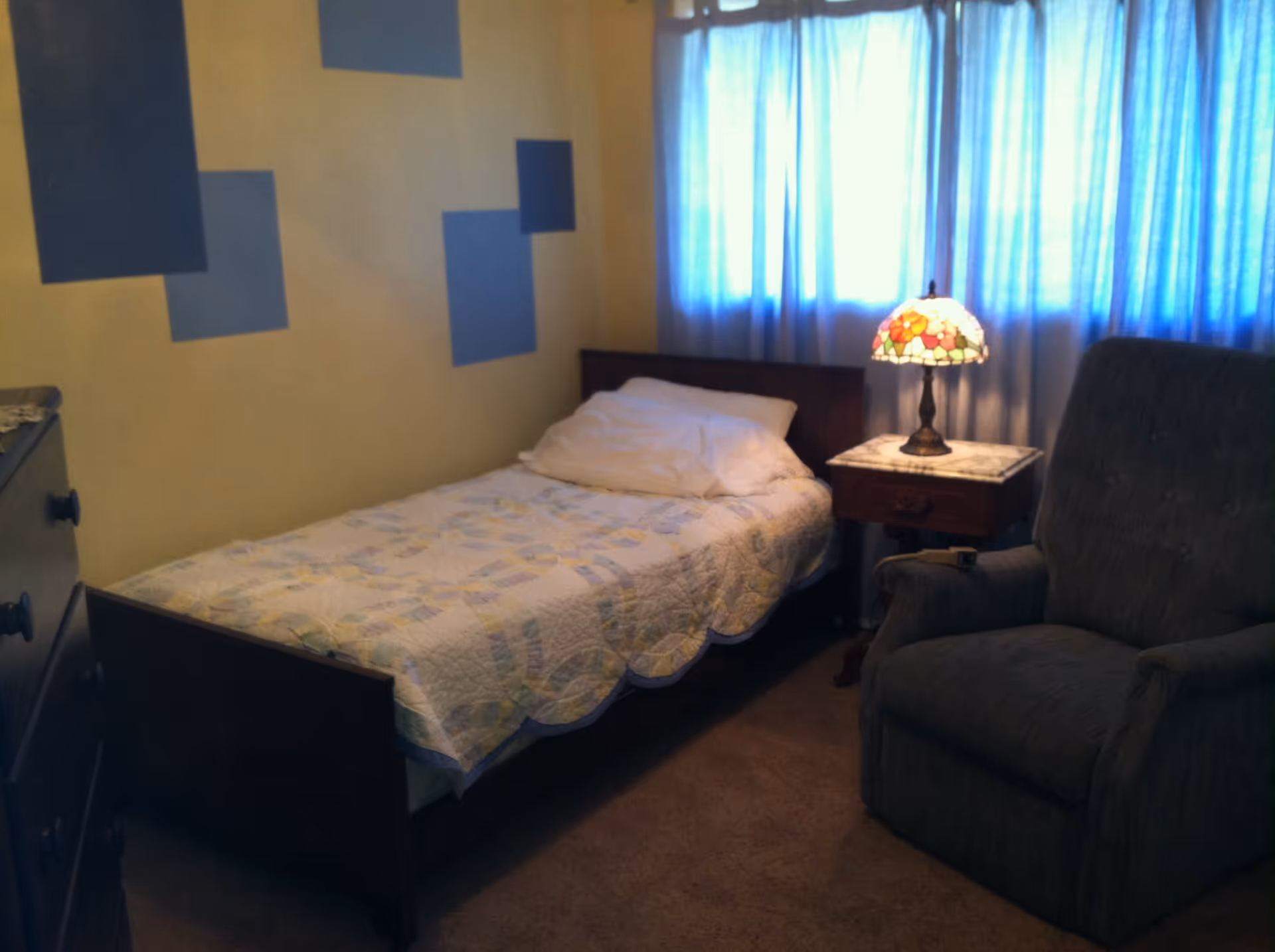 A small bedroom with a single bed covered with a patterned quilt and a pillow. Next to the bed is a wooden nightstand with a decorative lamp featuring a stained glass floral shade. A large upholstered armchair is positioned near the nightstand. The room has beige walls with blue square decorations and a window with blue curtains letting in light.