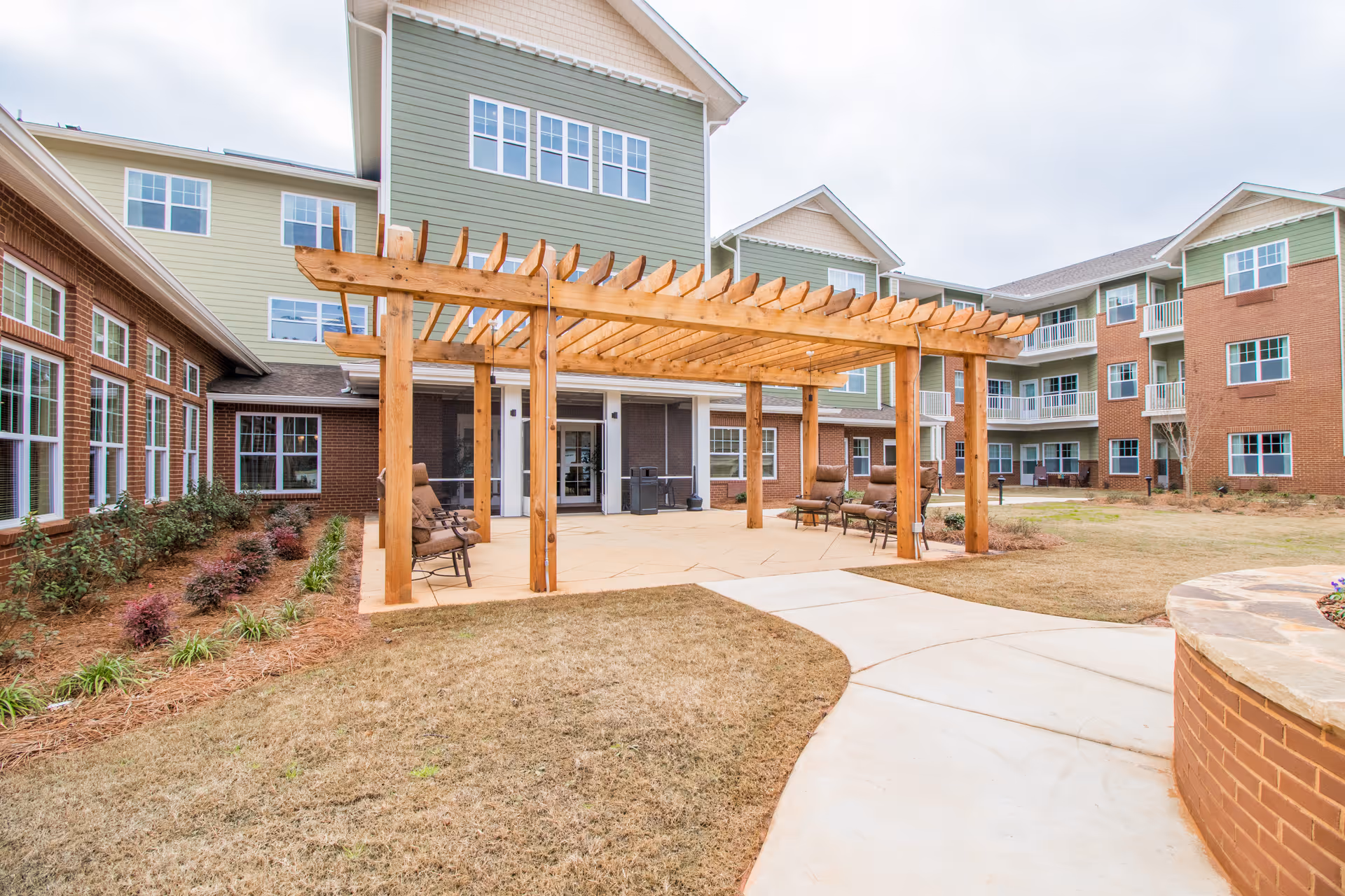 Outdoor patio area at Linwood Estates Gracious Retirement Living featuring a wooden pergola with several cushioned chairs underneath. The patio is surrounded by a well-maintained lawn and landscaping, with a multi-story building in the background that has green and brick exterior walls and multiple windows.