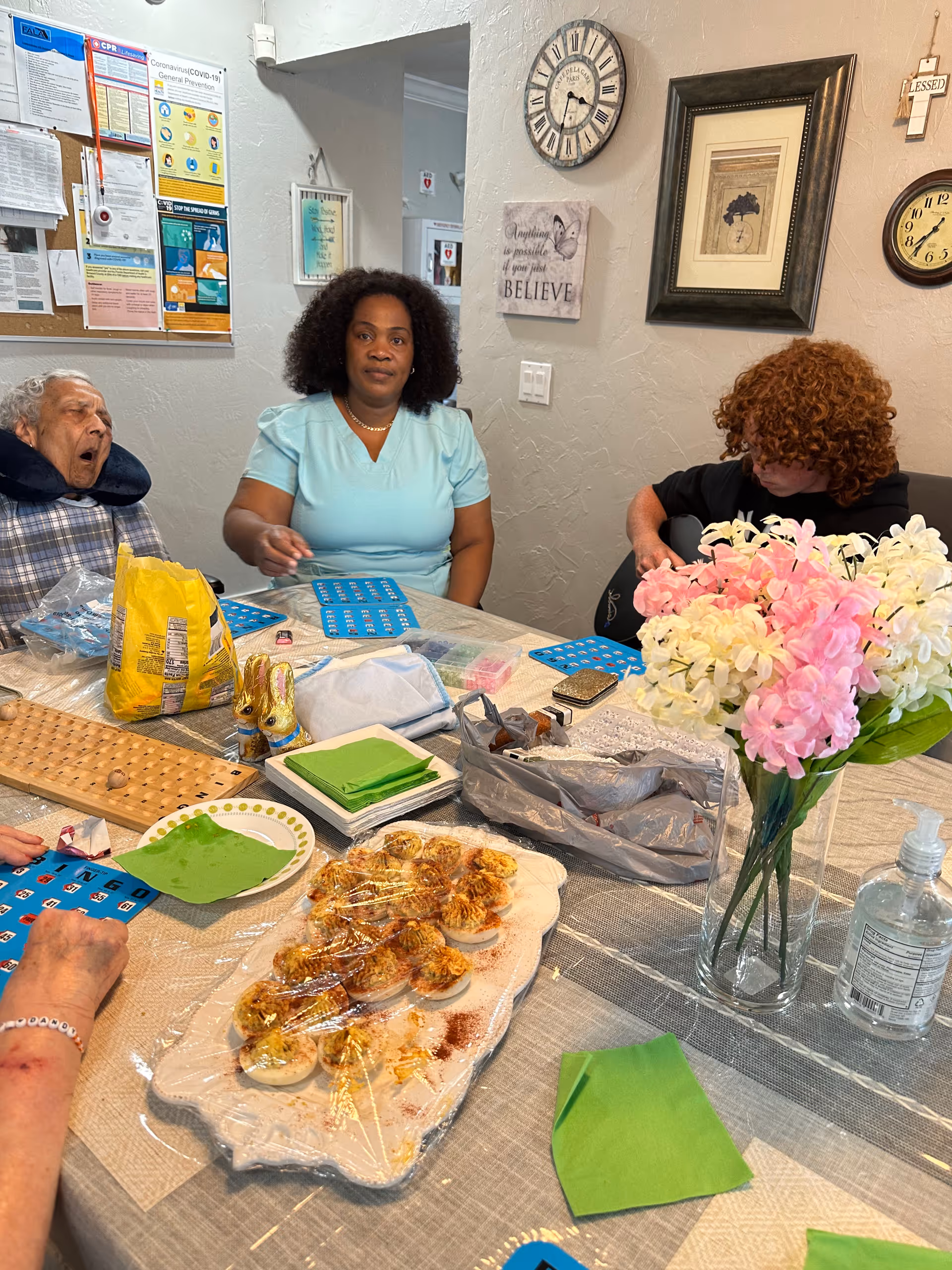 Residents and a staff member sit around a table covered with bingo cards, snacks, napkins and a vase of flowers in an assisted living common/dining room.