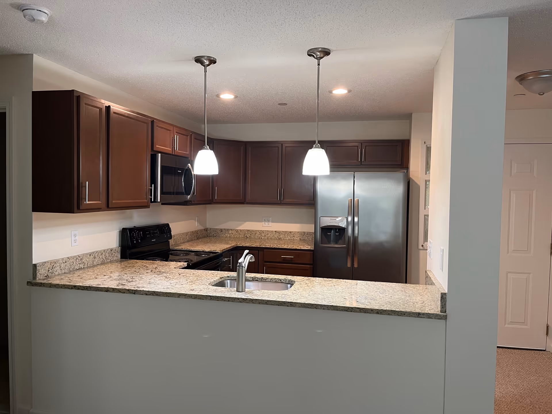 Modern kitchen with granite countertops, dark wood cabinets, stainless steel refrigerator, black stove, microwave, and two pendant lights hanging from the ceiling.
