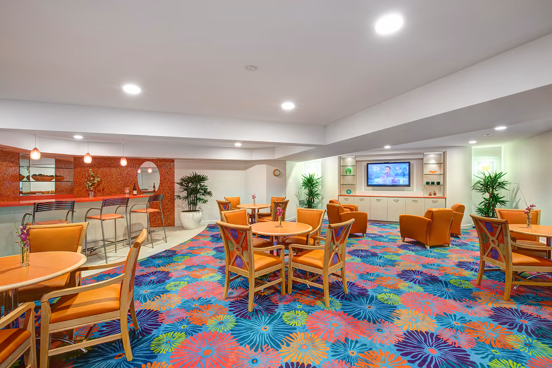 A brightly lit common area with colorful floral carpet, several round tables with orange chairs, a bar counter with high stools, potted plants, and a wall-mounted television.