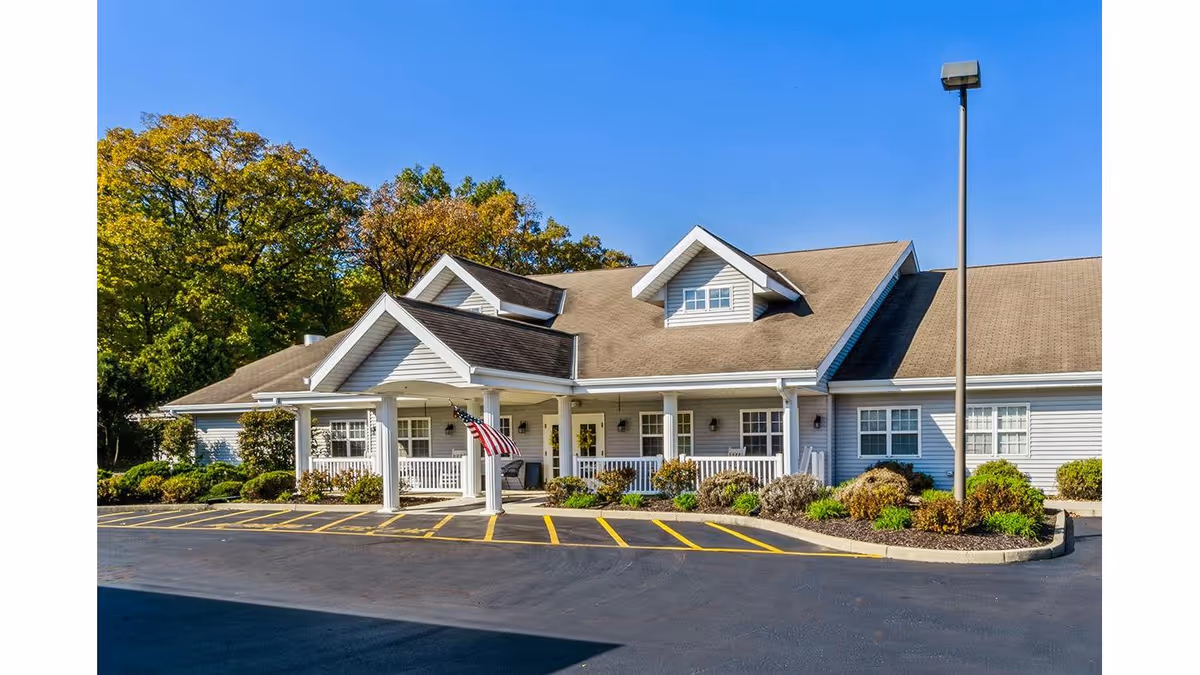 Single-story light-gray building with a covered front entrance, American flag, landscaping, and a parking lot under a clear blue sky.