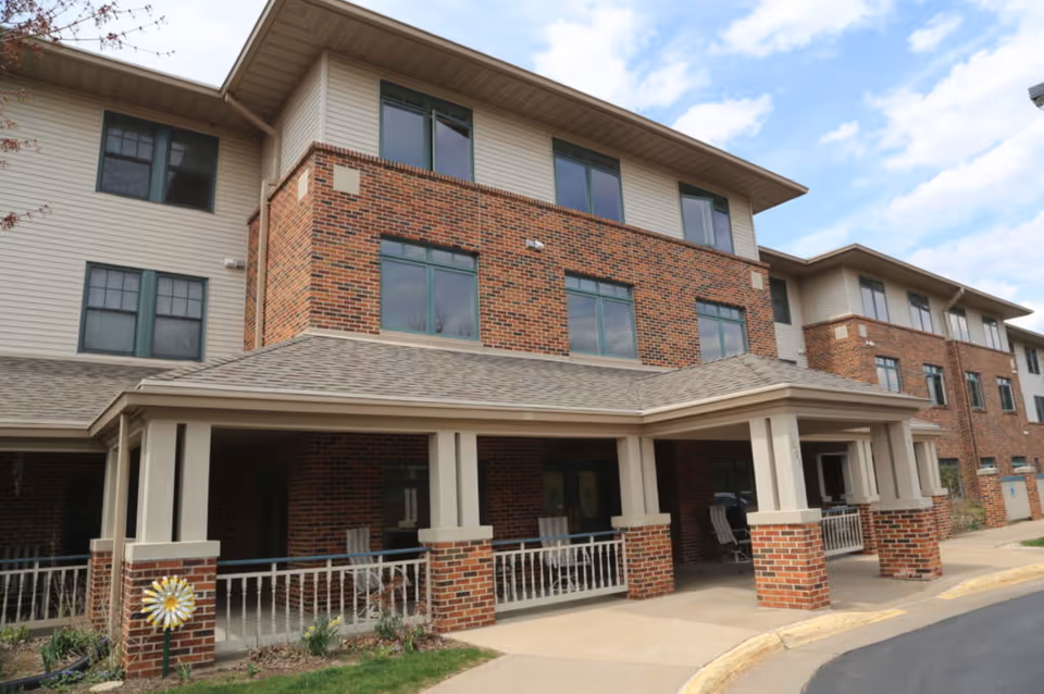 Exterior view of a three-story senior living facility building with a covered entrance supported by brick and beige columns. The building features a combination of red brick and beige siding with multiple windows. There is a small garden area with a decorative pinwheel near the entrance and a paved driveway in front.
