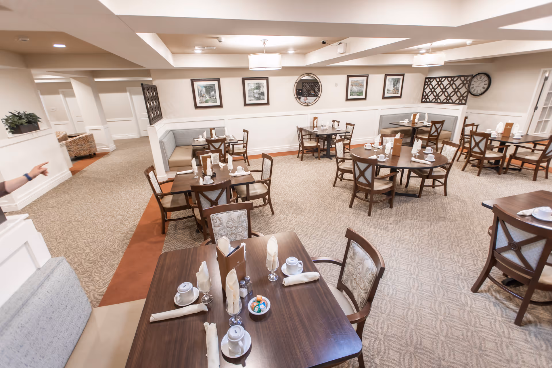 A dining room in a senior living facility with multiple wooden tables and chairs arranged neatly. Each table is set with white cups, napkins, and menus. The room has beige walls adorned with framed pictures and a round mirror. The floor is carpeted with a patterned design, and there are cushioned bench seats along the walls.