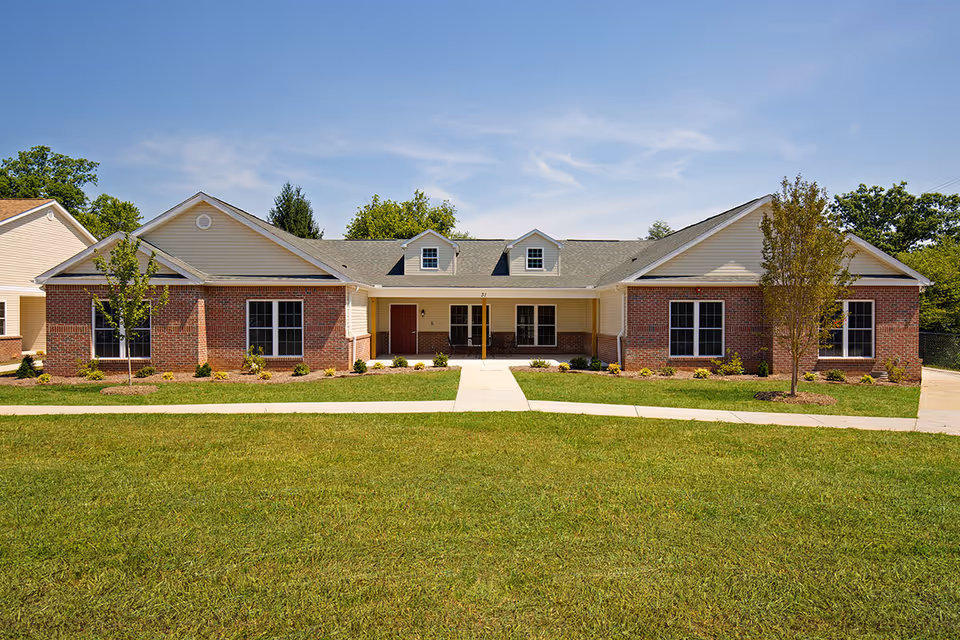 Single-story brick and siding building with a central entrance, two dormer windows on the roof, surrounded by a well-maintained lawn and small trees under a clear blue sky.