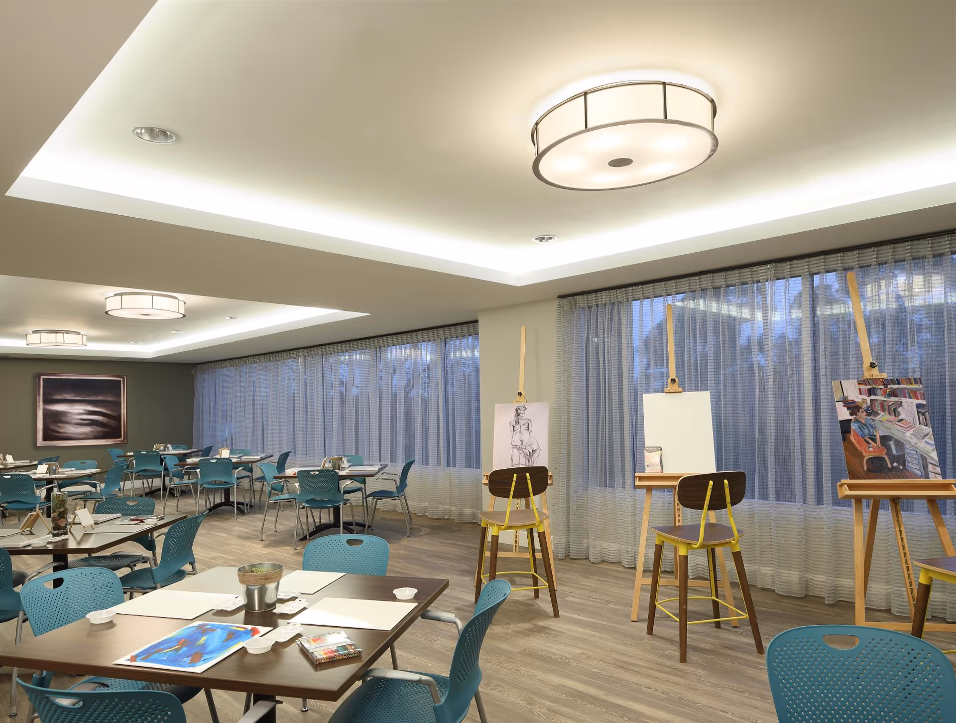 A spacious, well-lit room with multiple tables and blue chairs arranged for group activities. The room features large windows with sheer curtains, modern ceiling lights, and three easels with artwork and blank canvases, suggesting an art or activity room setting.