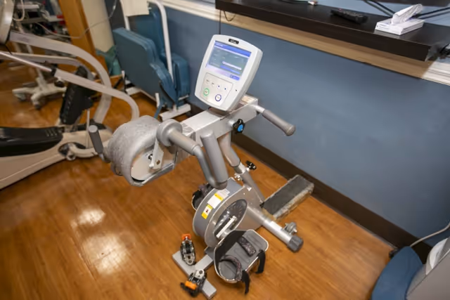 A physical therapy exercise machine with padded supports and digital display, placed on a wooden floor in a room with blue walls and a shelf holding tissues and other items.