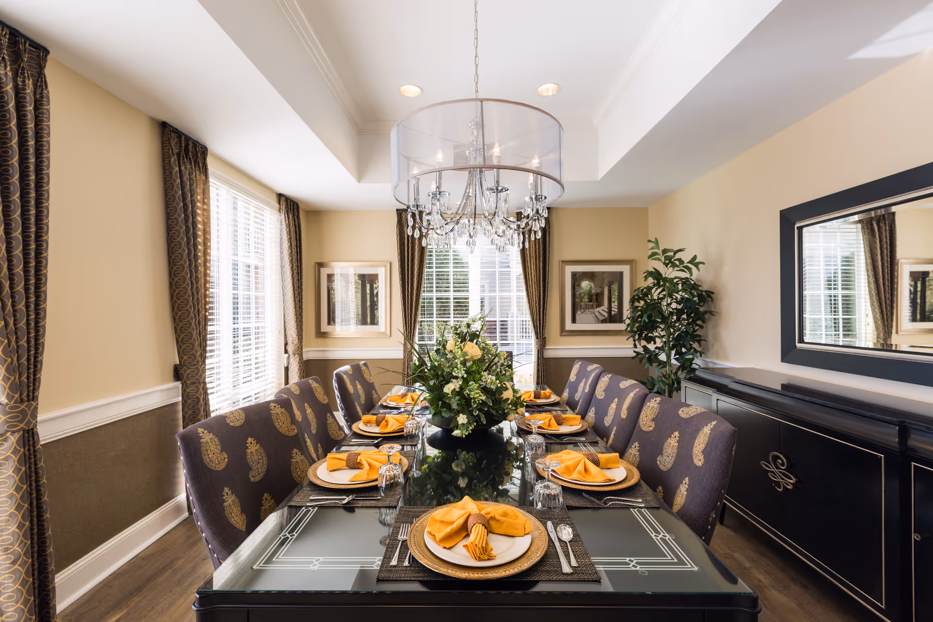 A formal dining room with a long glass-top table set for eight people. Each place setting has a white plate on a gold charger, a yellow folded napkin with a napkin ring, silverware, and an upside-down glass. The chairs are upholstered in a dark fabric with a gold pattern. The room has beige walls, large windows with patterned curtains, a crystal chandelier with a sheer drum shade, framed artwork, a large mirror, a black sideboard, and a green potted plant.