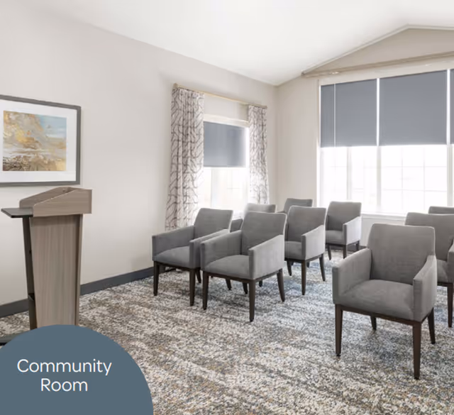 A community room with several gray upholstered armchairs arranged in rows facing a wooden podium. The room has light-colored walls, patterned curtains, and large windows with gray roller blinds partially drawn.