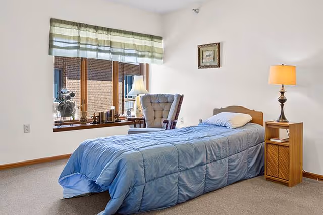 A simple bedroom with a single bed covered in a blue comforter and a white pillow. Next to the bed is a wooden nightstand with a lamp on it. There is a cushioned armchair near a large window with a green and white striped valance. The window sill has decorative items including a lamp and small plants. The walls are white and the floor is carpeted.