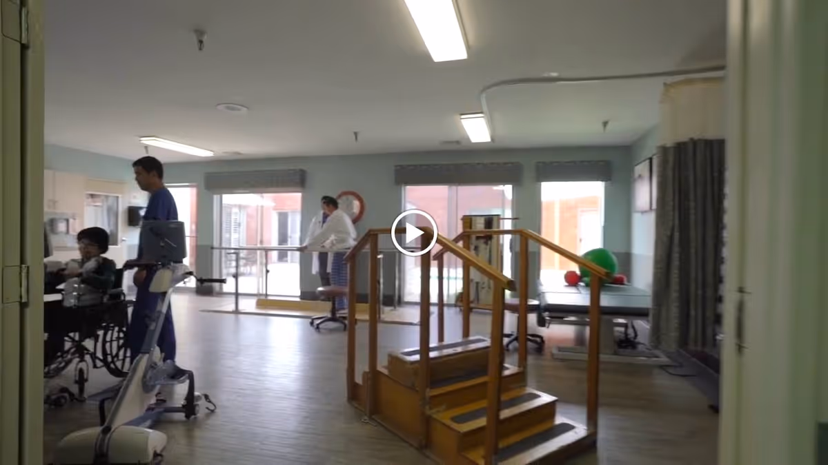 A rehabilitation therapy room with wooden stairs for physical therapy in the center, parallel bars near large windows, exercise balls on a table, and two caregivers assisting a person in a wheelchair.
