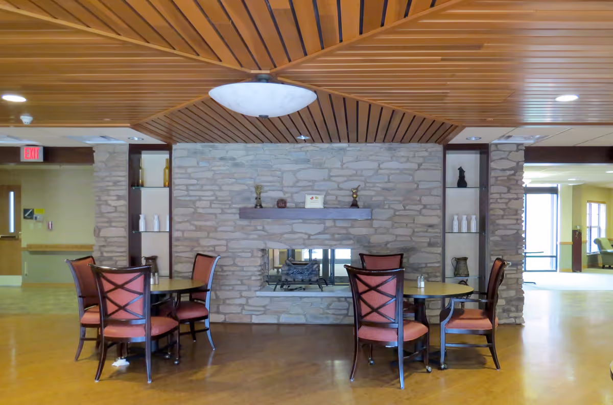 A cozy seating area in a senior living facility featuring two round tables each surrounded by four wooden chairs with red cushions. The background shows a stone wall with a built-in fireplace and decorative items on a wooden mantel. The ceiling has a wooden slat design with a large round light fixture in the center. The room has a warm and inviting atmosphere with visible hallways and exit signs in the distance.