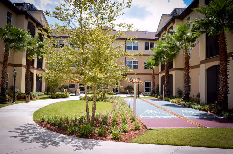 Outdoor courtyard area at Hawthorne Estates of Ocala featuring a shuffleboard court, palm trees, a central tree with surrounding plants, and a curved walkway between two beige multi-story buildings under a partly cloudy sky.