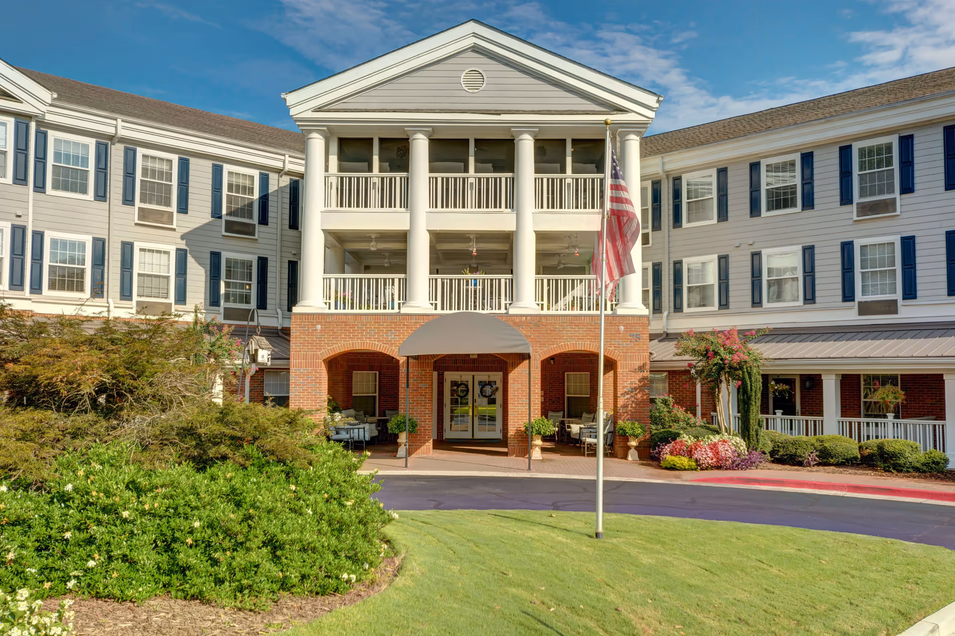 Front exterior view of Historic Roswell Place, a multi-story senior living facility with white siding, blue shutters, and a brick entrance. The building features a covered porch with columns, balconies, an American flag on a pole in front, and well-maintained landscaping with bushes and flowers.