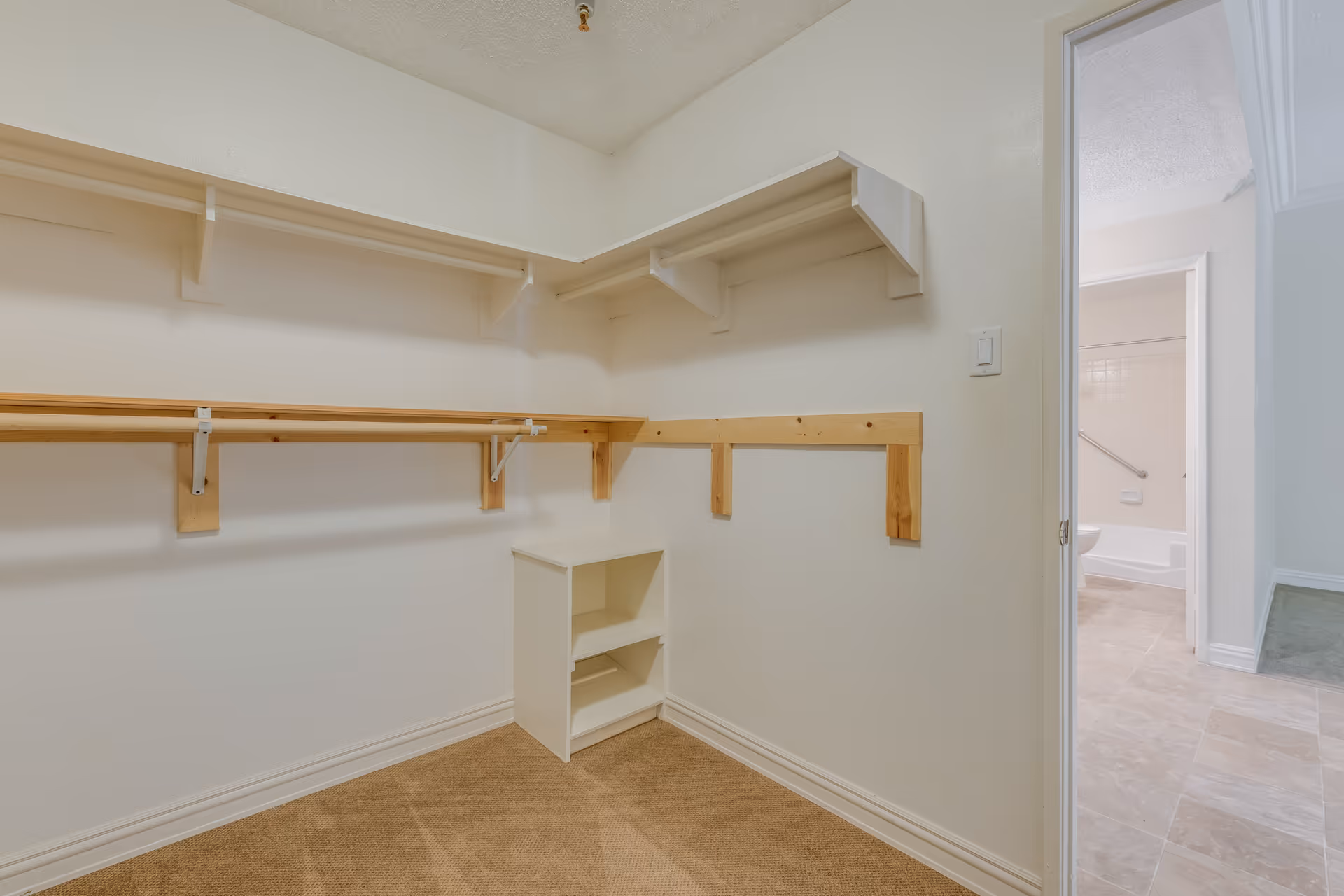 Empty walk-in closet with beige carpet, white walls, wooden hanging rods, and white shelves. The closet opens into a hallway leading to a bathroom with a bathtub and tiled floor.